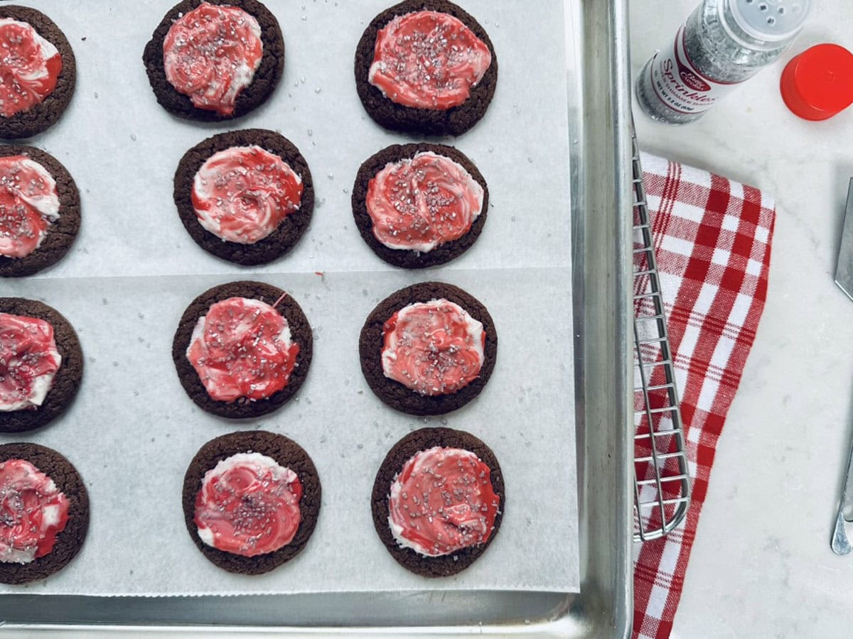 Chocolate cookies with white and red swirled frosting and sprinkles are arranged on a parchment-lined baking sheet. A red and white checkered cloth and a shaker are nearby on the counter.