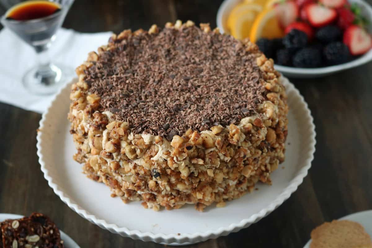 A round cake covered in chopped nuts on the sides and topped with chocolate shavings, displayed on a white cake stand. In the background, there is a fruit plate and a glass with a dark drink.