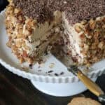 A layered cake with creamy frosting, chopped nuts on the sides, and chocolate shavings on top sits on a white cake stand. A slice has been cut, revealing the inner layers, with a knife beside the cake.