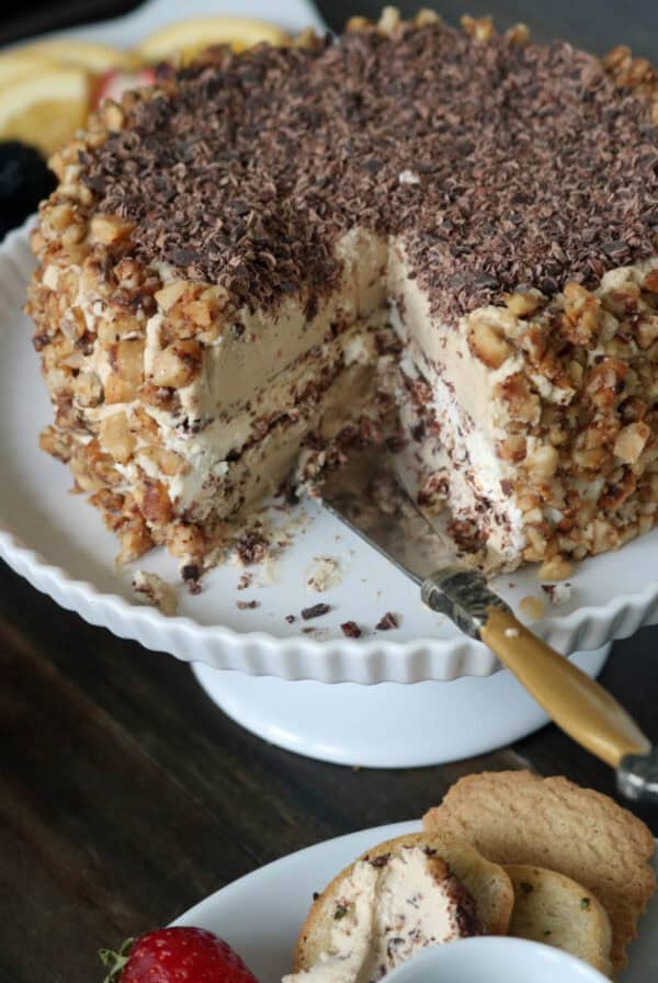 A layered cake with creamy frosting, chopped nuts on the sides, and chocolate shavings on top sits on a white cake stand. A slice has been cut, revealing the inner layers, with a knife beside the cake.