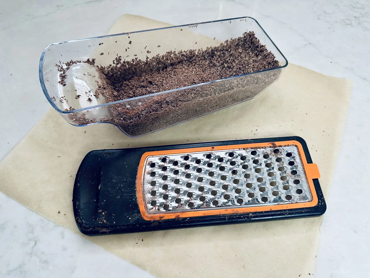 A box grater with grated chocolate inside and some chocolate shavings scattered around, placed on a sheet of parchment paper on a light countertop.