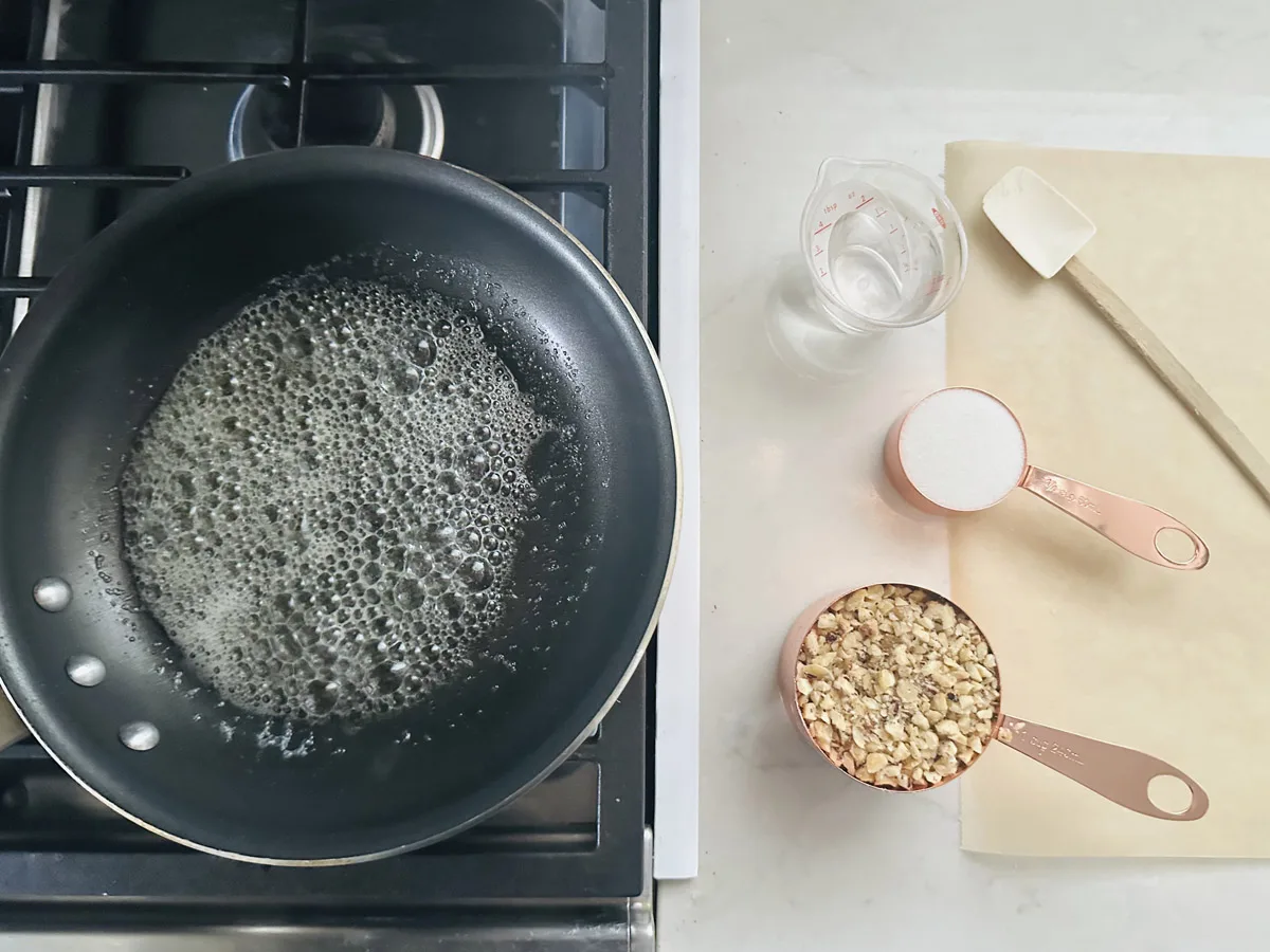 A frying pan with bubbling liquid sits on a stove. Next to it are a measuring cup of water, a spatula, a cup of chopped nuts, and a cup of sugar on a white countertop with parchment paper.