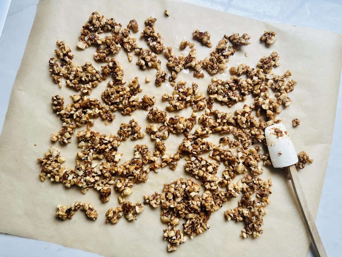 Clusters of nut brittle are scattered on a sheet of parchment paper, with a white spatula resting next to them. The brittle appears golden brown and chunky, suggesting a homemade mixture.