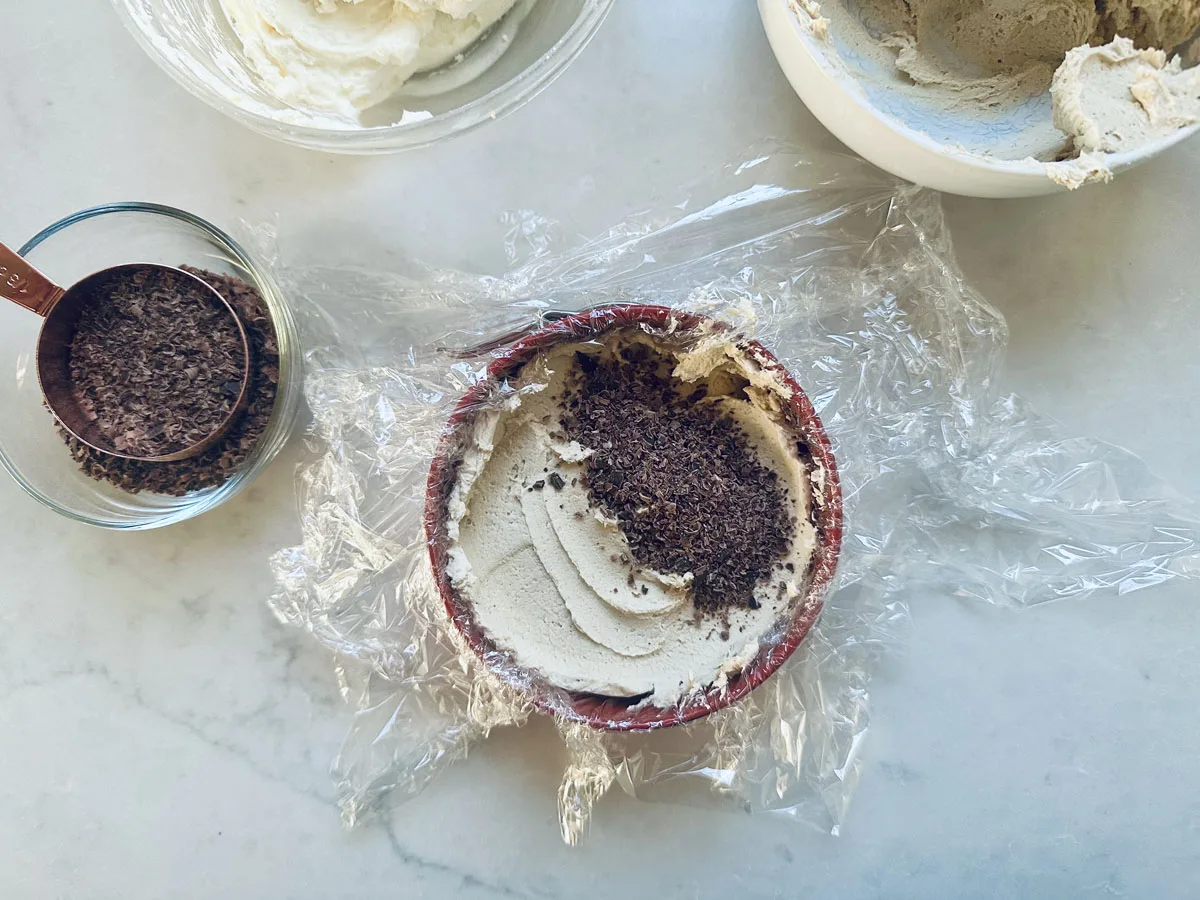 A bowl lined with plastic wrap contains a creamy mixture topped with grated chocolate. Nearby are a small glass bowl with more chocolate shavings and another bowl with leftover cream on a light countertop.