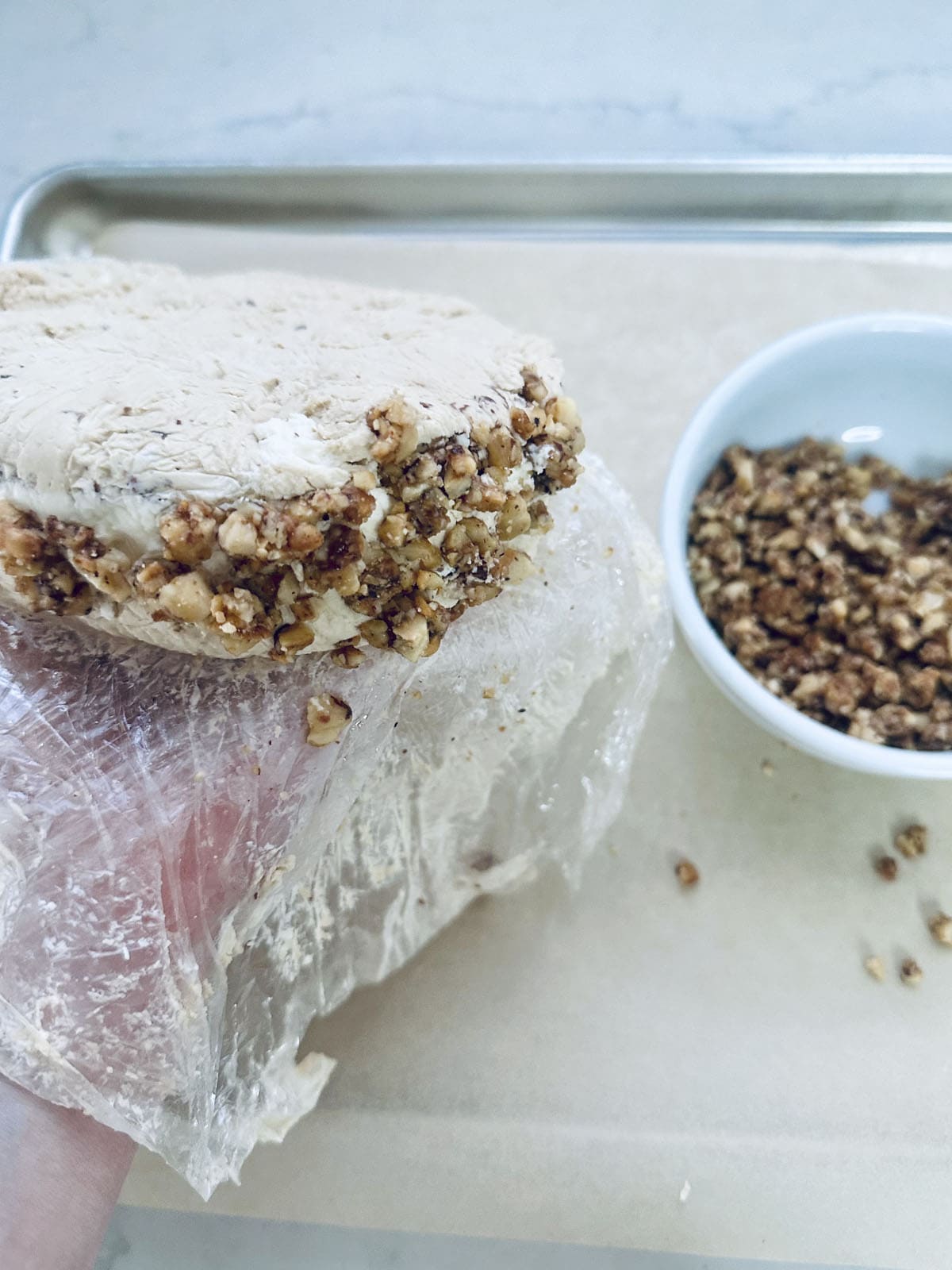 A hand holds a block of dough partially wrapped in plastic, pressing its edge into chopped nuts on a baking sheet. A small white bowl filled with nuts sits nearby.