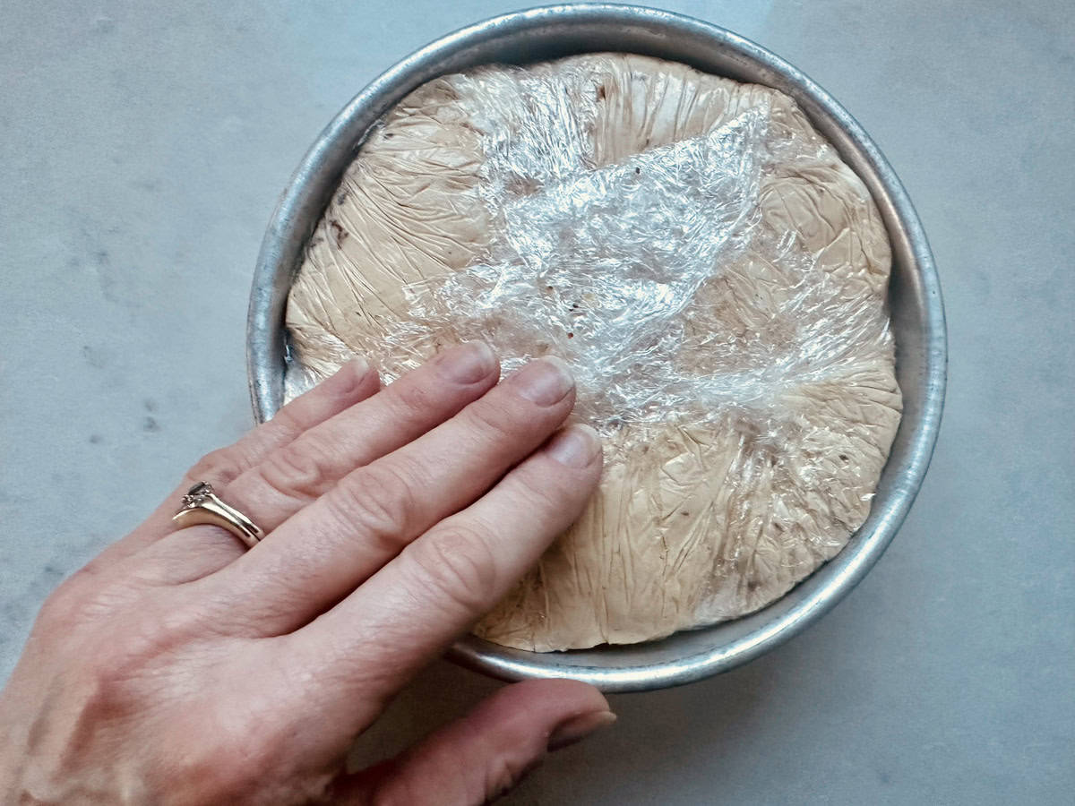A hand with a ring presses plastic wrap over dough rising in a round metal pan on a light-colored surface.