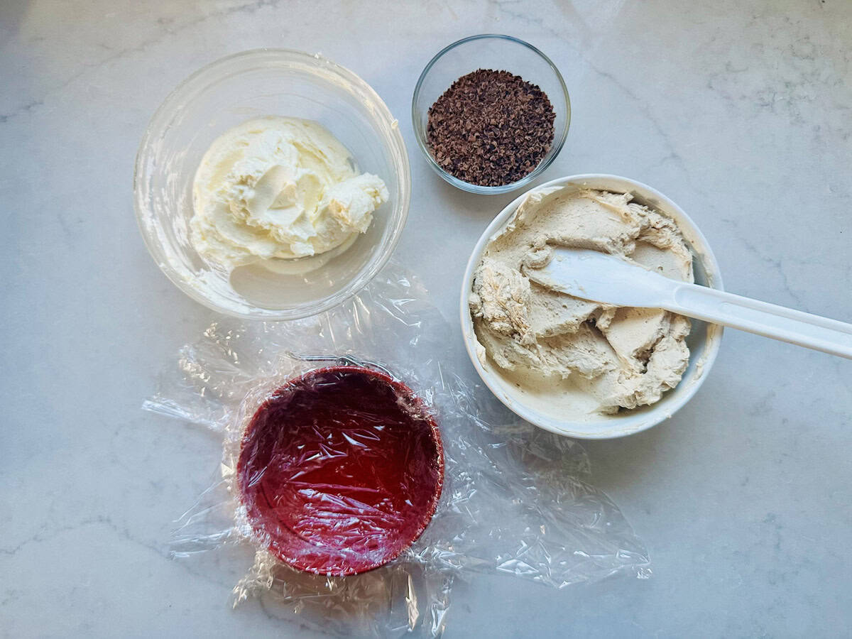 A countertop displays a bowl of whipped cream, a bowl of coffee ice cream with a spatula, a small bowl of ground coffee, and a red bowl lined with plastic wrap.