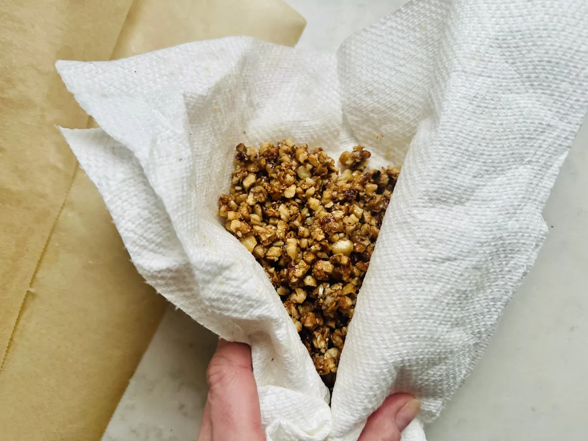 A hand holds a paper towel containing a mixture of finely chopped, caramelized nuts or seeds, with parchment paper visible nearby on a light surface.