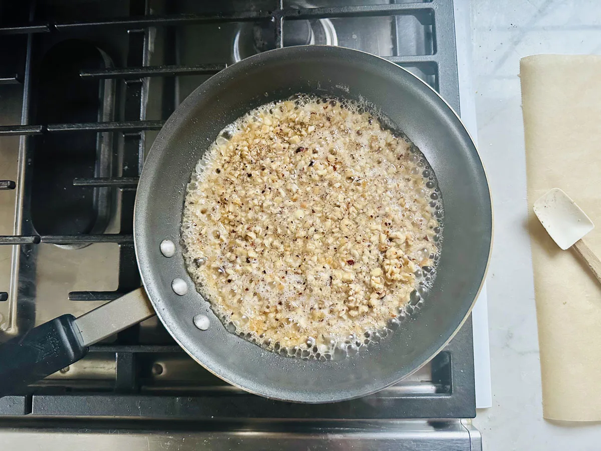 A frying pan on a stovetop contains bubbling, chopped nuts or seeds being toasted in a liquid, with a spatula and parchment paper nearby on a marble counter.