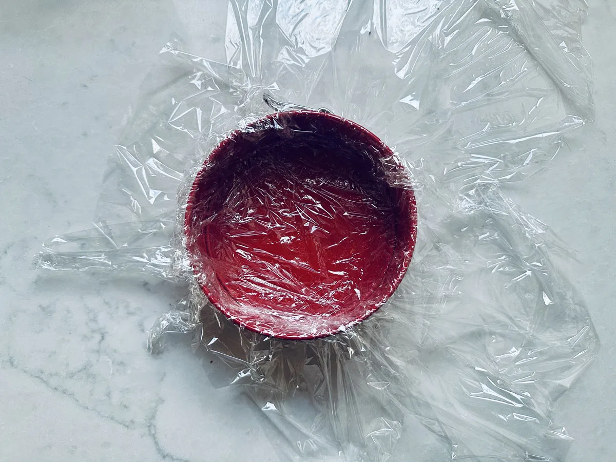 A red bowl covered with clear plastic wrap sits on a light-colored, marbled surface. The plastic wrap is spread out messily around the bowl.