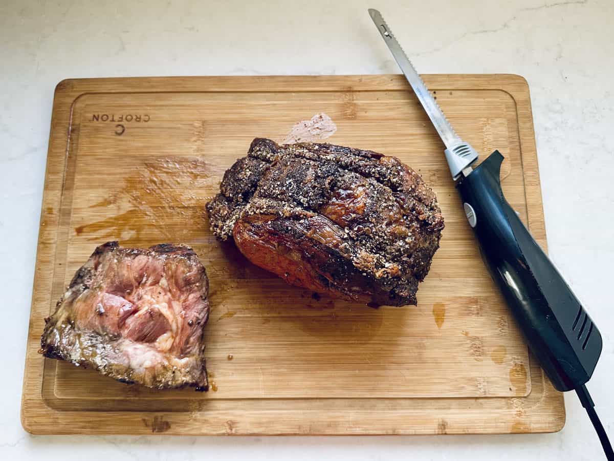 A Standing Rib Roast with Au Jus sits on a wooden cutting board beside a sliced portion of meat and an electric carving knife, all on a light-colored countertop.