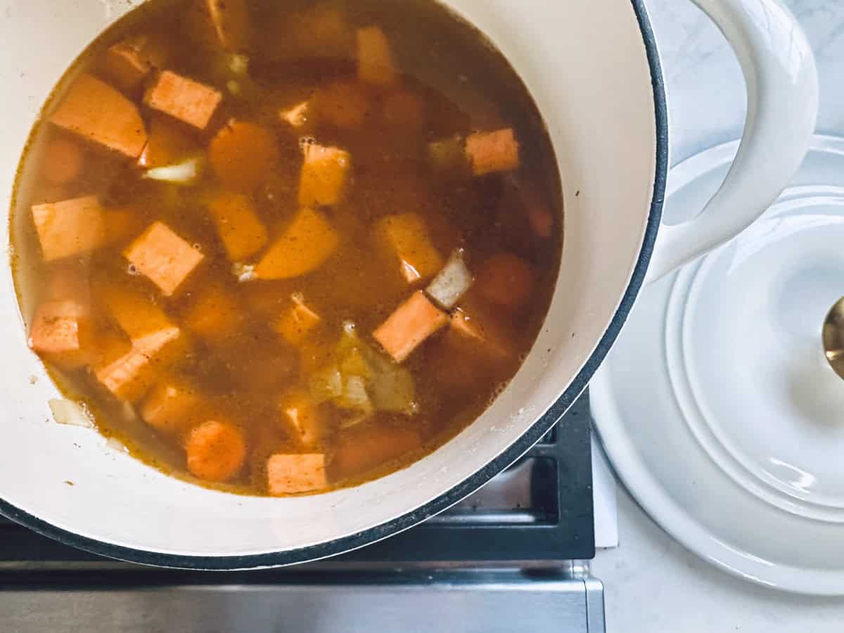 A white pot on a stove contains a simmering broth with chunks of orange sweet potatoes and diced onions. The pot lid rests nearby on the counter.