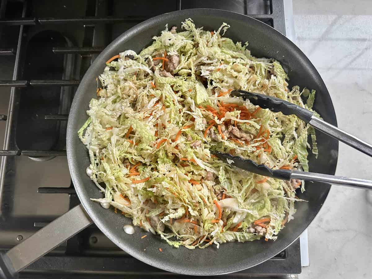 A frying pan on a stove filled with shredded cabbage, carrots, and ground meat being sautéed. Metal tongs rest on the pan, ready to stir the mixture as it cooks.