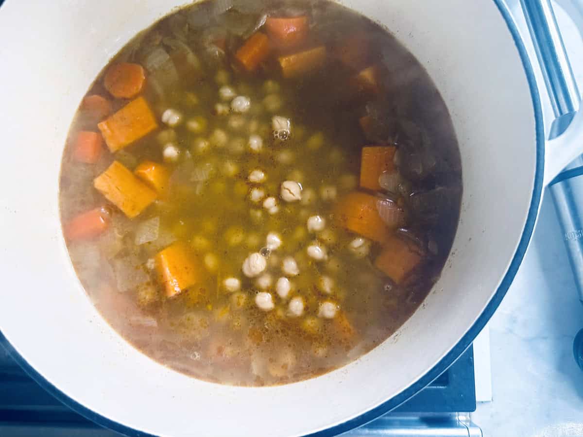A pot of soup with chickpeas, chopped carrots, and onions simmering in broth on a stovetop.