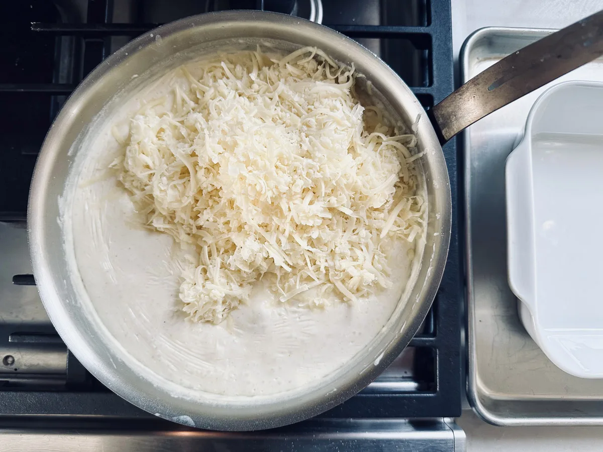 A metal pan on a stove contains a creamy white sauce topped with a generous pile of shredded cheese, ready to be mixed in. A baking dish sits nearby on a metal tray.
