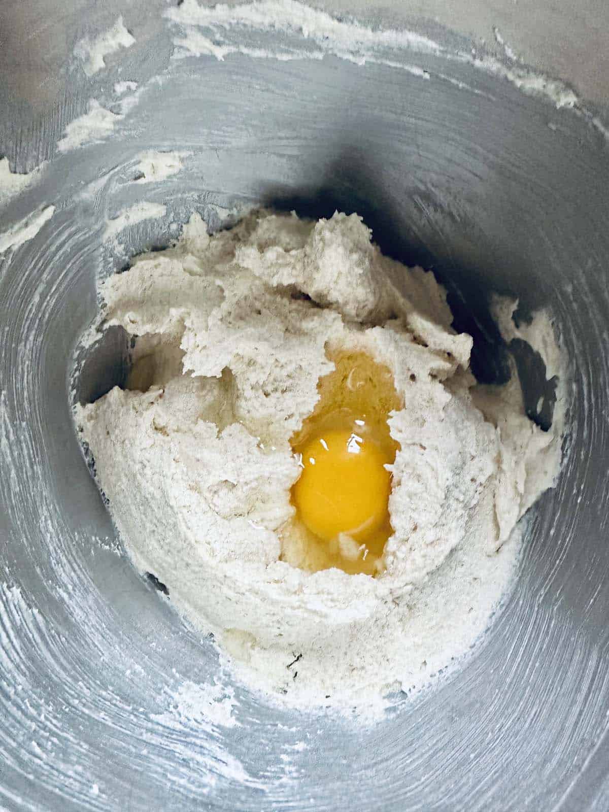 A cracked raw egg sits in the center of a mound of flour and dough mixture inside a metal mixing bowl, ready to be blended together.