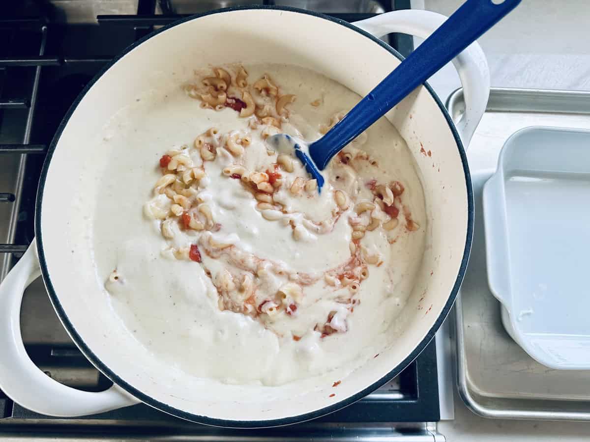 A white pot on a stove contains pasta being mixed into a creamy white sauce with bits of tomato, using a blue spatula. A white baking dish sits nearby on a metal tray.