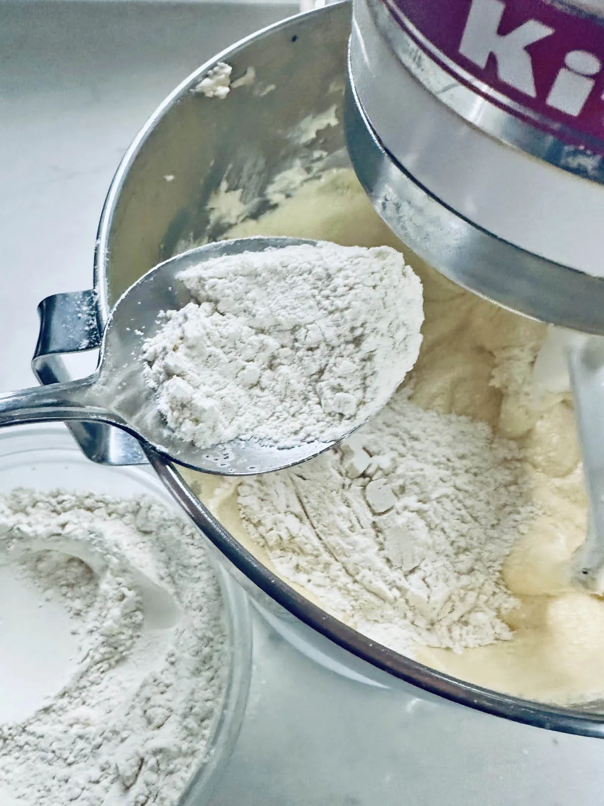 A spoonful of flour is being added to a mixer bowl containing dough, with a stand mixer and a separate bowl of flour visible nearby.
