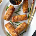 A white plate with five golden-brown egg rolls, garnished with green onion slices. A small bowl of dipping sauce and a pair of chopsticks are placed beside the egg rolls.