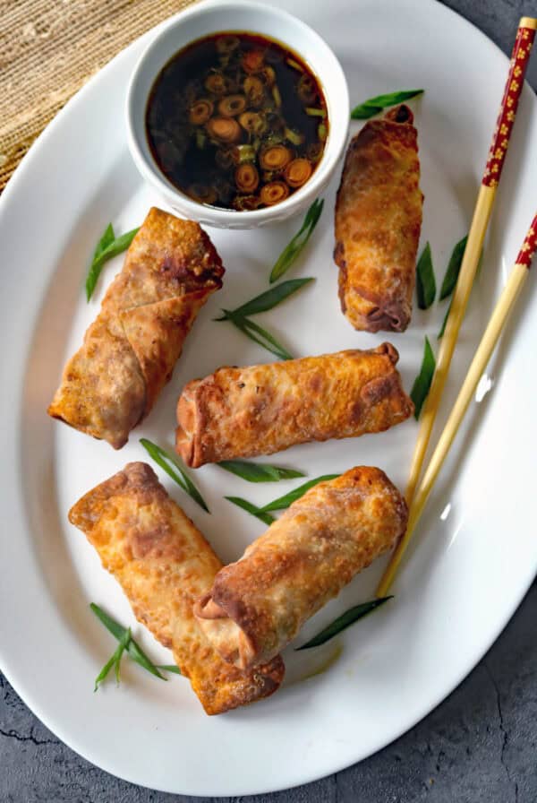 A white plate with five golden-brown egg rolls, garnished with green onion slices. A small bowl of dipping sauce and a pair of chopsticks are placed beside the egg rolls.