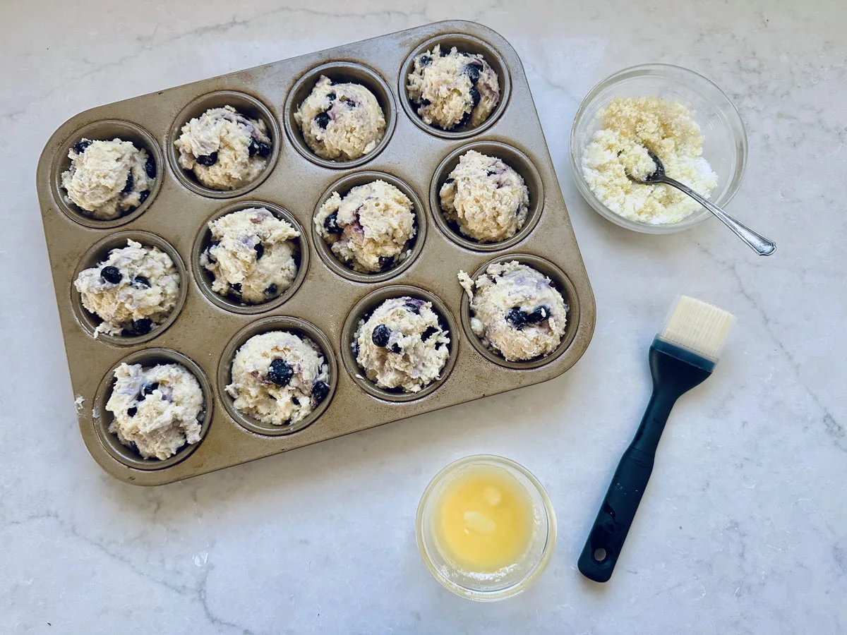 A muffin tin filled with unbaked blueberry muffin batter sits on a marble countertop. Nearby are a bowl of crumb topping, a pastry brush, and a small bowl of melted butter.