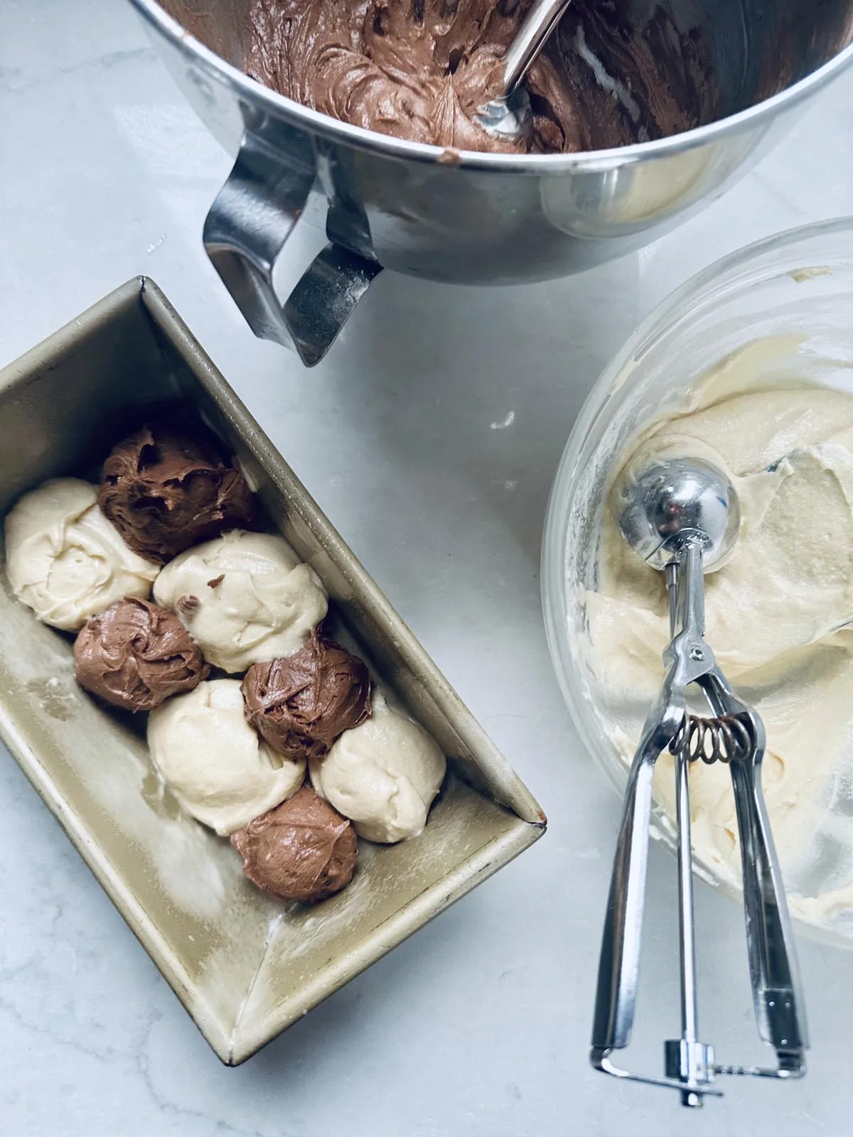 A loaf pan with scoops of chocolate and vanilla batter, a metal ice cream scoop, a bowl with vanilla batter, and a mixing bowl with chocolate batter on a light countertop.