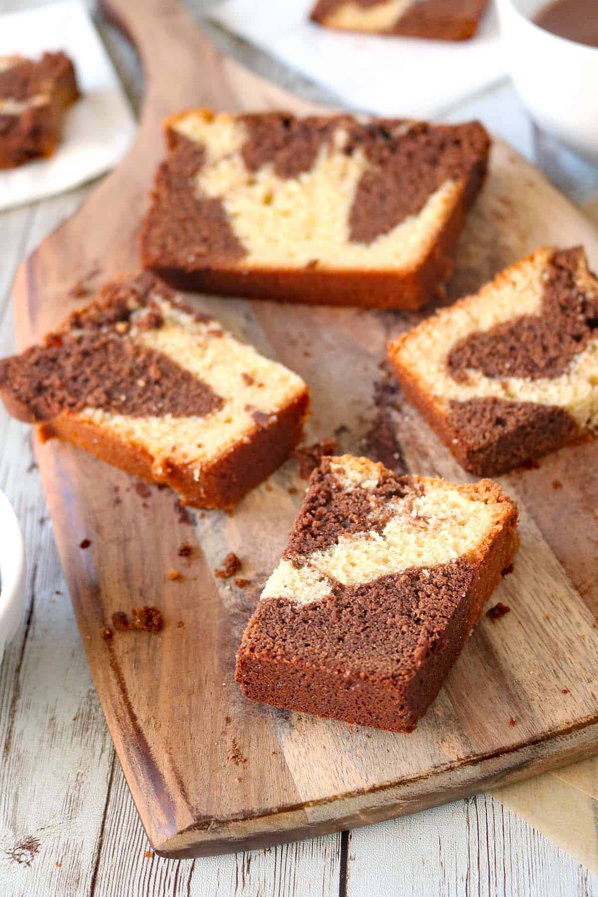 Four slices of marble cake with swirled chocolate and vanilla patterns are arranged on a wooden serving board, with cake crumbs scattered around on a rustic wooden surface.
