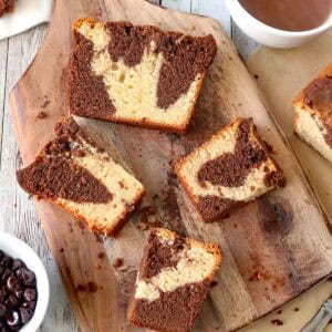 Slices of marble cake with swirls of chocolate and vanilla rest on a wooden board, surrounded by a bowl of chocolate chips and a cup of hot chocolate on a rustic white wooden table.