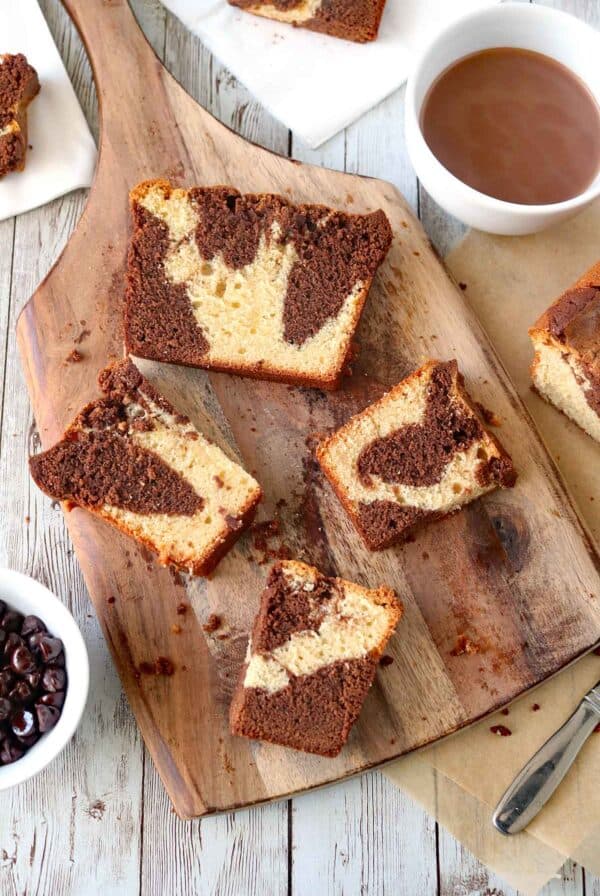 Slices of marble cake with swirls of chocolate and vanilla rest on a wooden board, surrounded by a bowl of chocolate chips and a cup of hot chocolate on a rustic white wooden table.