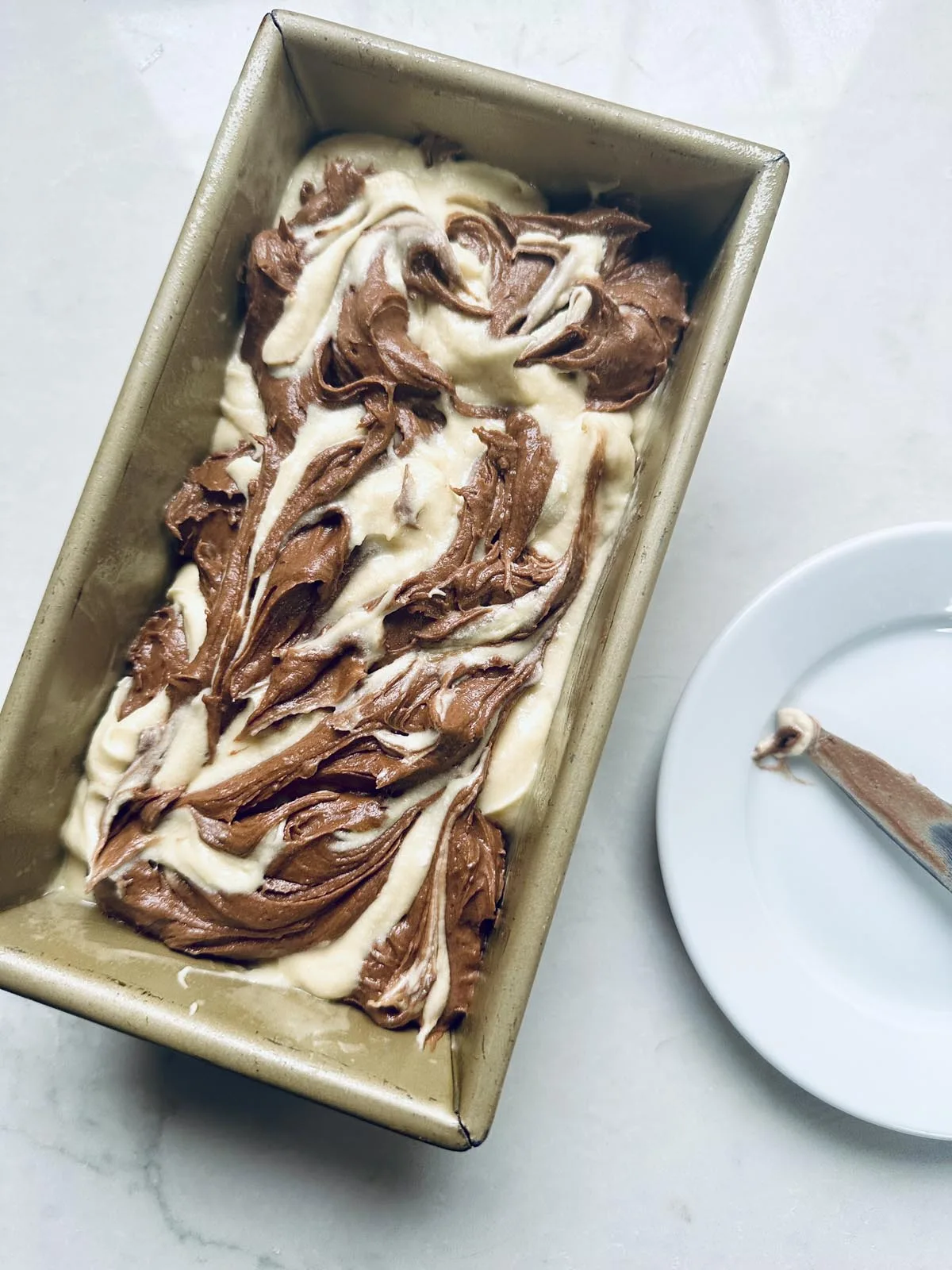A loaf pan filled with swirled chocolate and vanilla cake batter sits on a light surface next to a white plate with a spoon covered in batter.