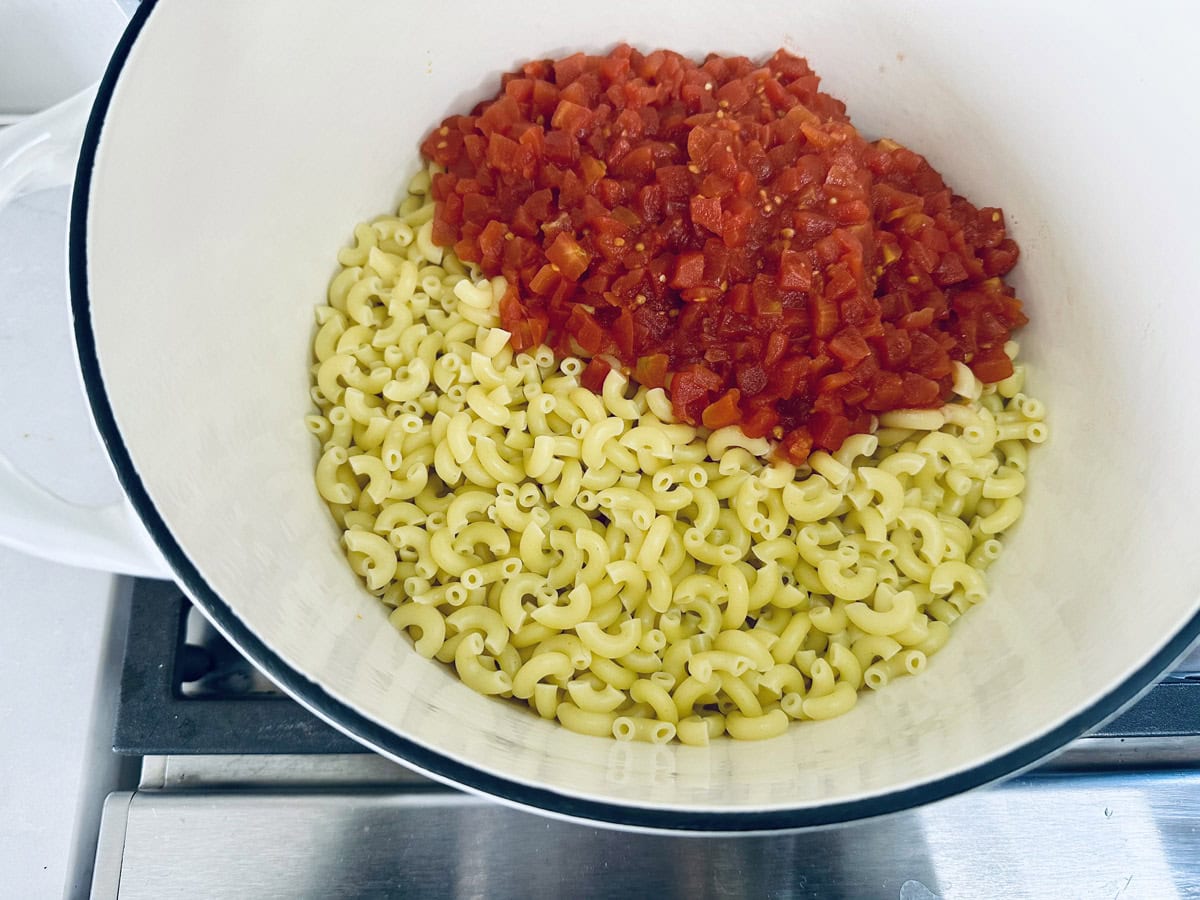 A white pot on a stove contains cooked elbow macaroni pasta and a pile of diced tomatoes, ready to be mixed together.
