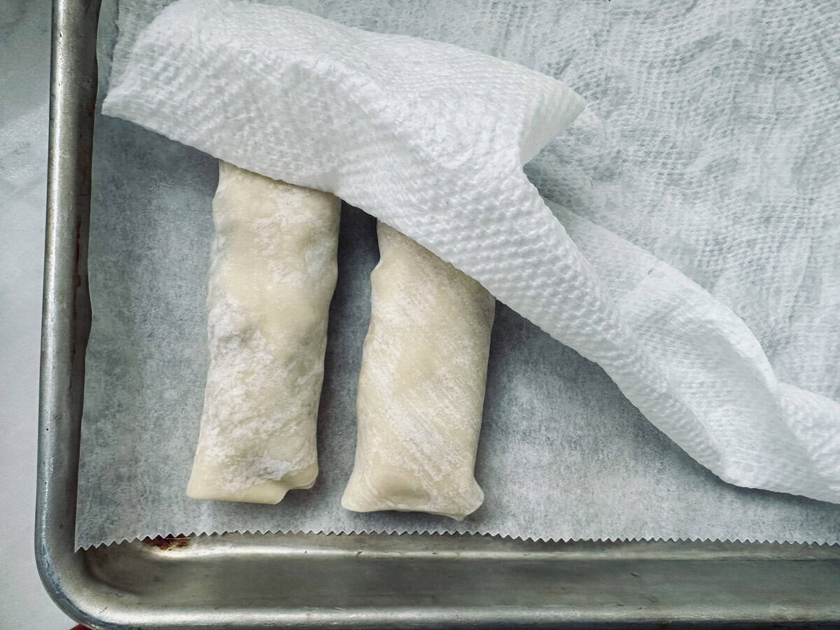 Two uncooked spring rolls covered with a damp paper towel rest on a parchment-lined metal baking sheet, preparing to prevent them from drying out.