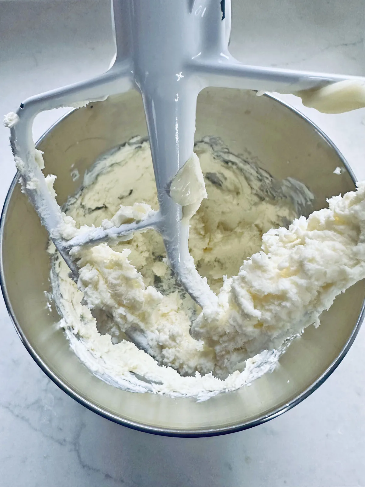 A close-up of a stand mixer with a paddle attachment mixing a creamy, pale batter in a metal bowl on a white countertop.