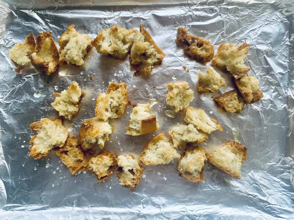 Chunks of torn bread scattered on a sheet of aluminum foil, lightly drizzled with oil and sprinkled with salt, ready for toasting or baking.