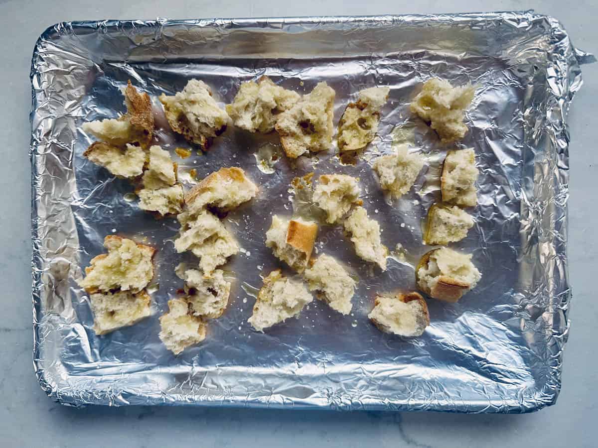 Chunks of torn bread scattered on a foil-lined baking tray, ready for toasting or baking. The bread appears partially crispy and is placed on a light-colored surface.