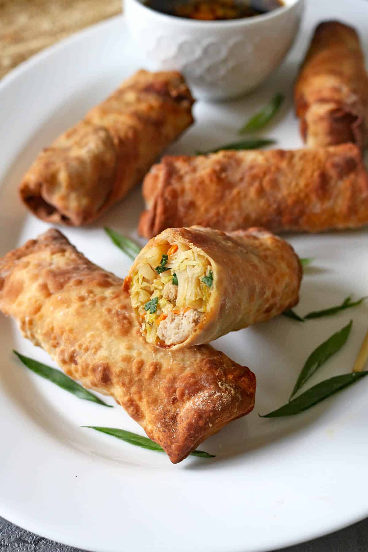 Four golden-brown egg rolls on a white plate, one cut open to show a filling of vegetables and meat, with green garnish and a bowl of dipping sauce in the background.