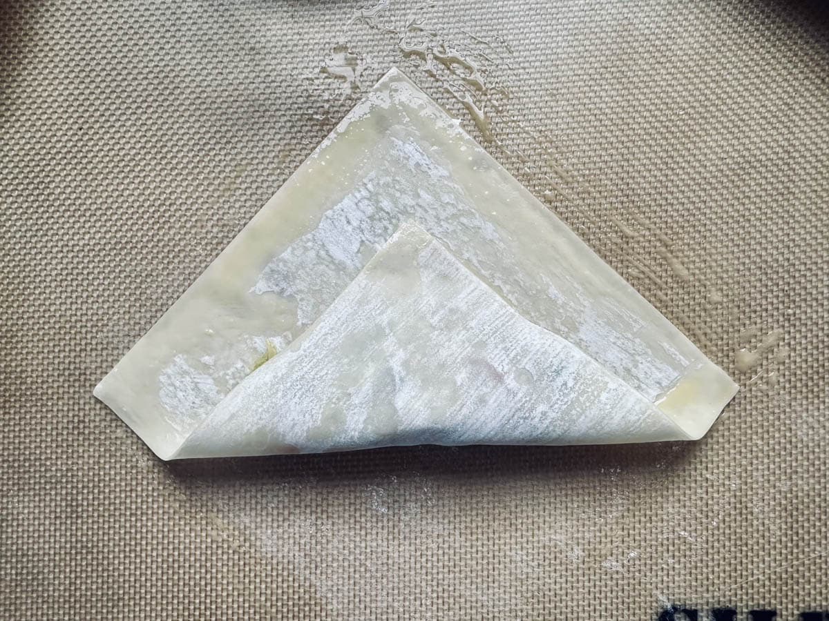 A single piece of dough is partially folded into a triangle shape on a brown silicone baking mat, ready for further preparation.