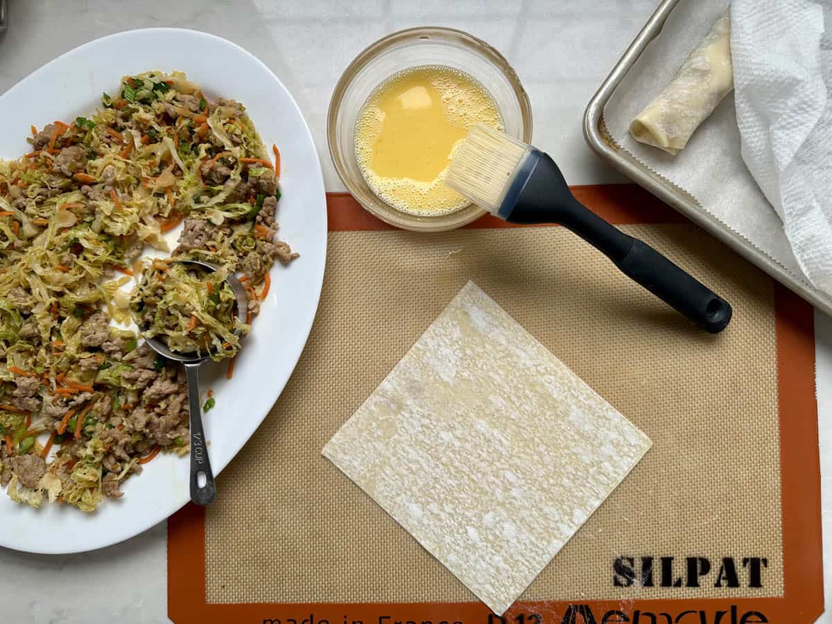 A plate of cooked meat and vegetable filling, a square wonton wrapper on a baking mat, a bowl of beaten egg with a brush, and a tray with a rolled wrapper are arranged on a kitchen counter.