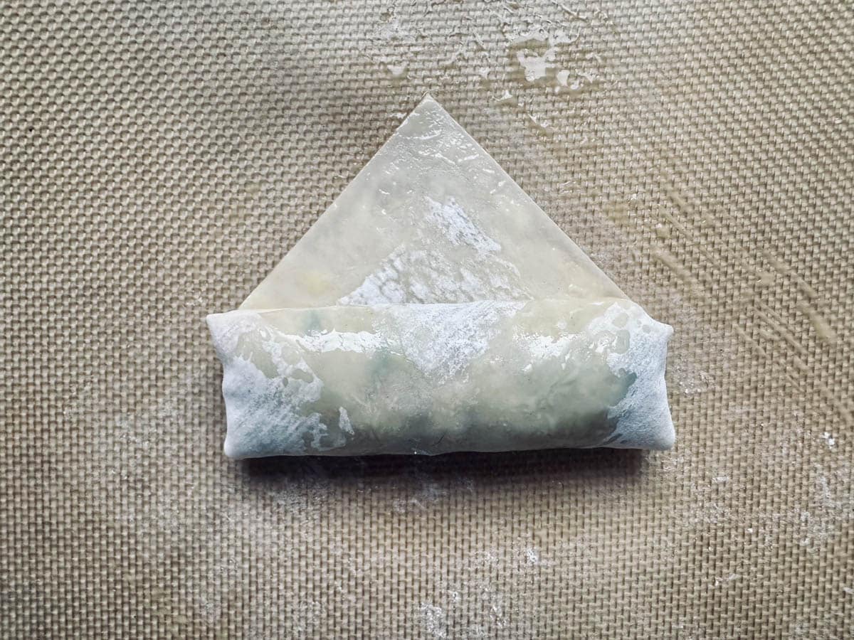 A close-up of a partially rolled spring roll with a translucent wrapper on a textured, light brown surface dusted with flour.
