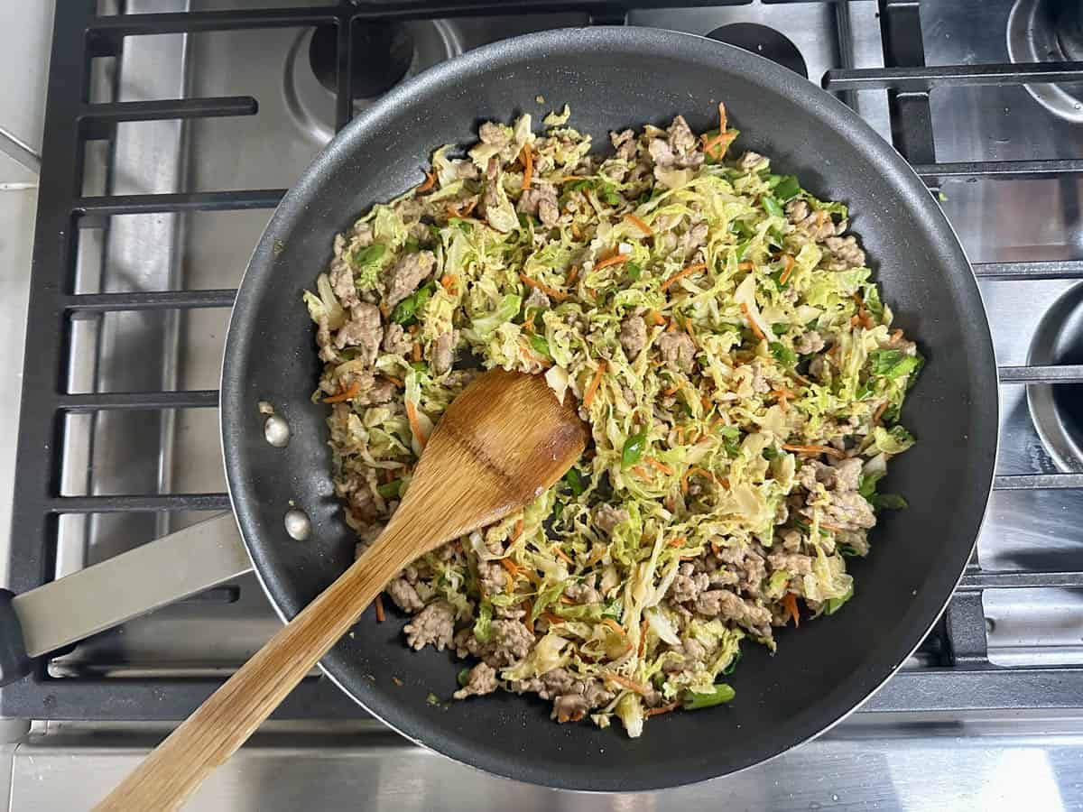 A skillet on a stove contains a stir-fry of ground meat, shredded cabbage, and carrots, with a wooden spatula resting inside the pan.