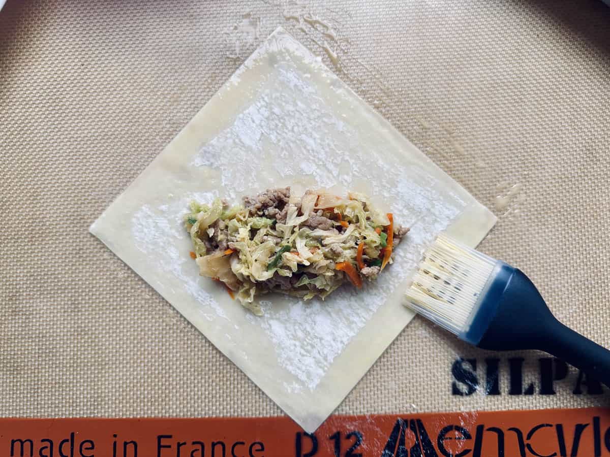 A square wrapper with a meat and vegetable filling is on a baking mat, next to a pastry brush. The filling includes shredded cabbage and carrots. The wrapper is dusted with flour, ready to be folded.