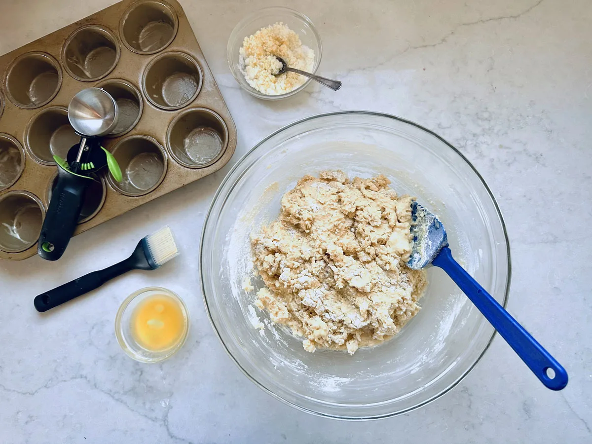 A glass bowl with biscuit dough and a blue spatula sits on a countertop, surrounded by a muffin tin, measuring tools, a pastry brush, a small bowl of egg wash, and a bowl of crumbled cheese.