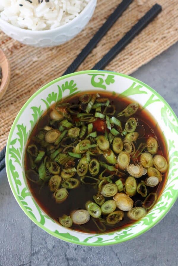 A bowl of dark soy sauce topped with sliced green onions, placed on a gray surface next to a bowl of white rice and a pair of black chopsticks.