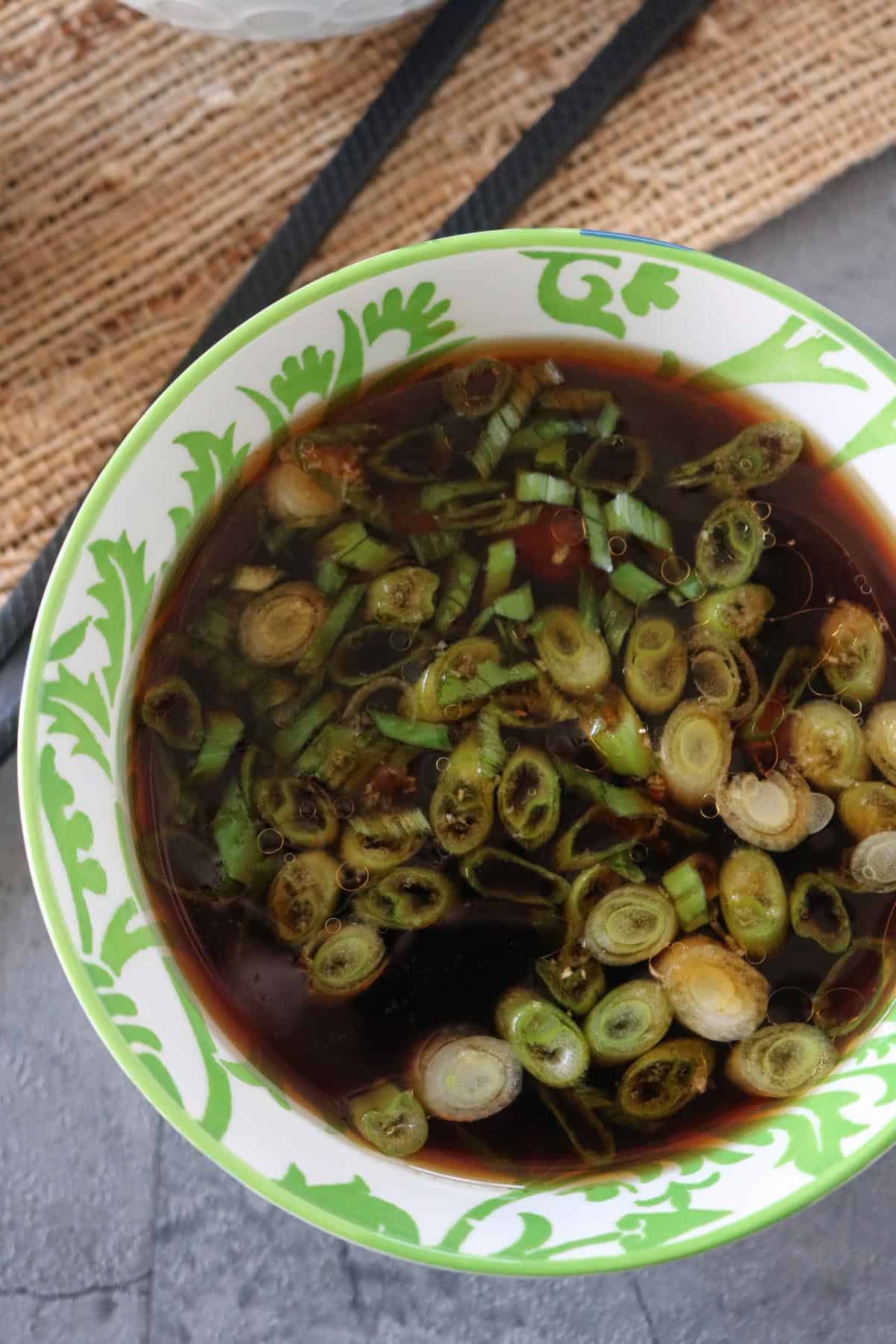 A bowl of dark soy-based dipping sauce topped with sliced green onions, in a white bowl with green decorative patterns, placed on a gray textured surface.
