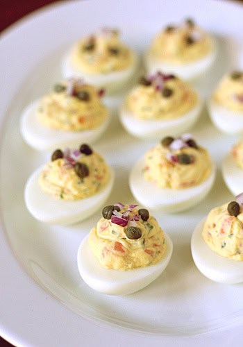 A plate of deviled eggs arranged neatly, each filled with a creamy yellow yolk mixture and topped with capers and finely chopped red onions.