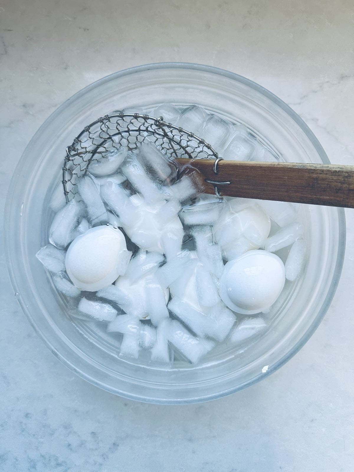 A wire skimmer rests in a glass bowl filled with ice water, ice cubes, and three white eggs, likely being cooled after boiling.