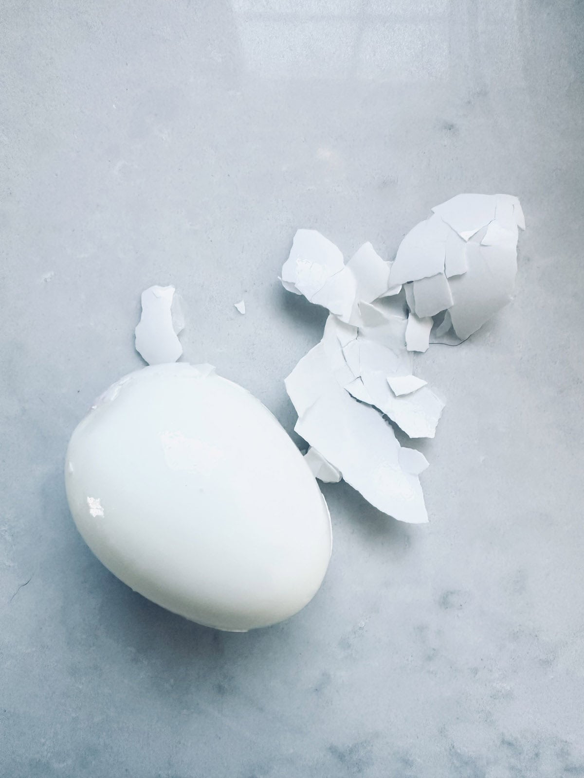 A peeled hard-boiled egg sits on a light gray surface next to pieces of its cracked eggshell.