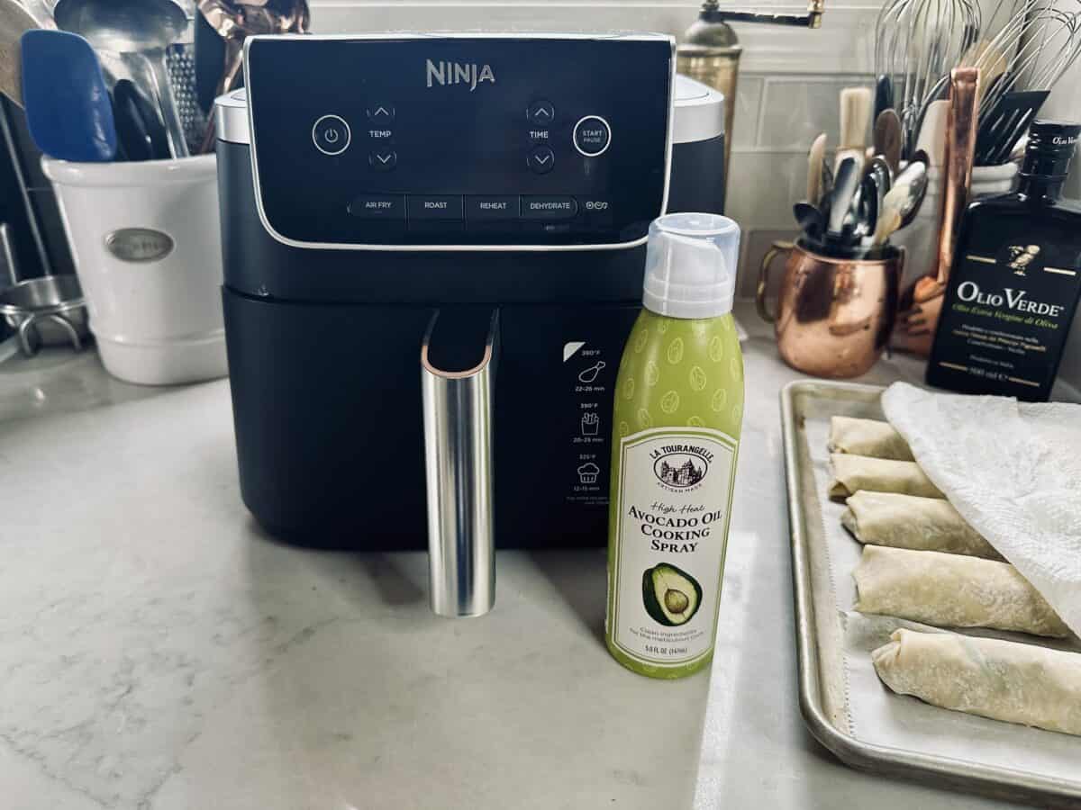 A Ninja air fryer sits on a kitchen counter next to a can of avocado oil cooking spray, with a tray of uncooked spring rolls covered by a paper towel nearby. Cooking utensils and olive oil are in the background.