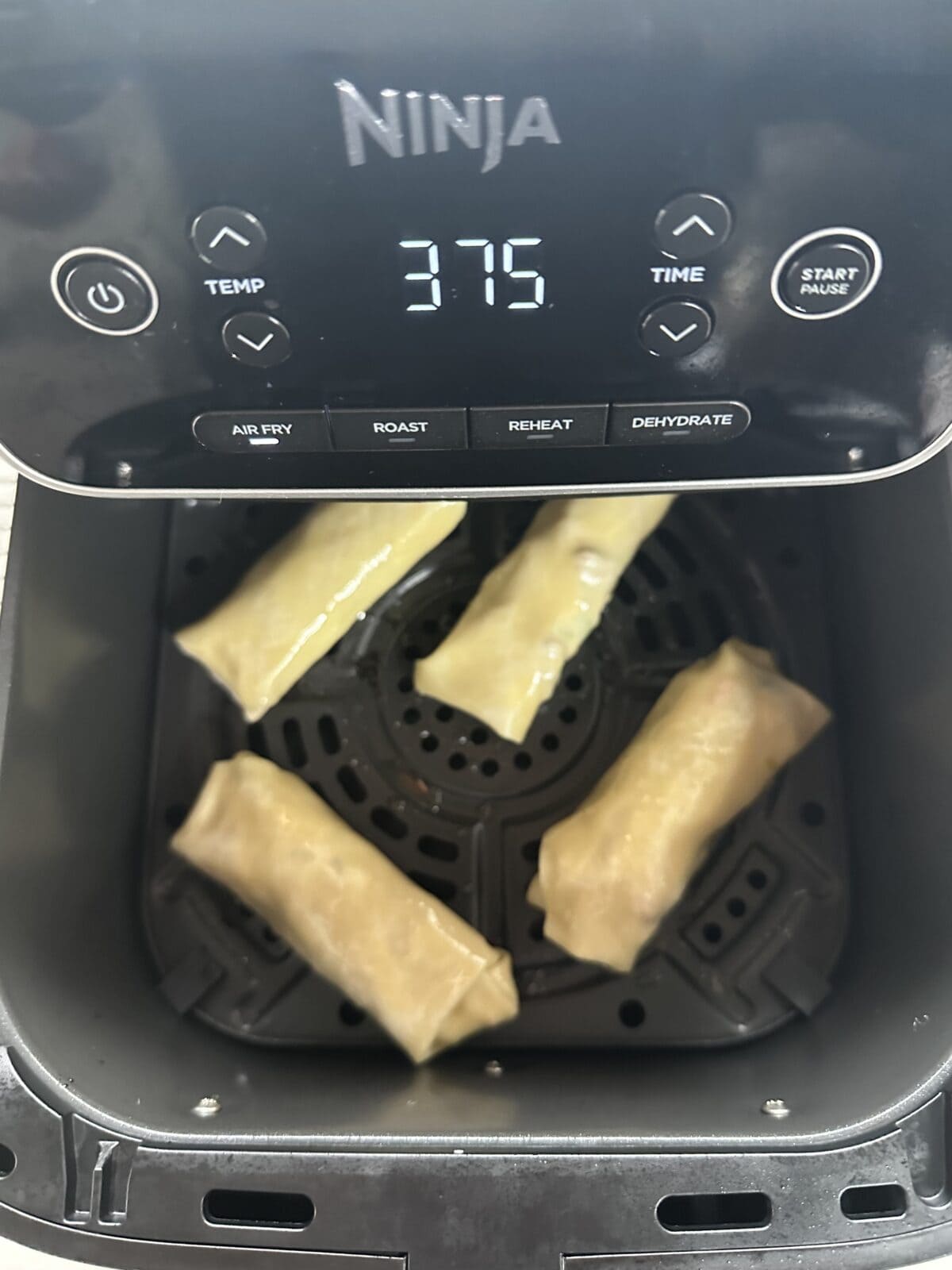 Four uncooked spring rolls are placed in the basket of a Ninja air fryer set to 375 degrees Fahrenheit, ready to be air fried. The control panel and basket interior are visible.