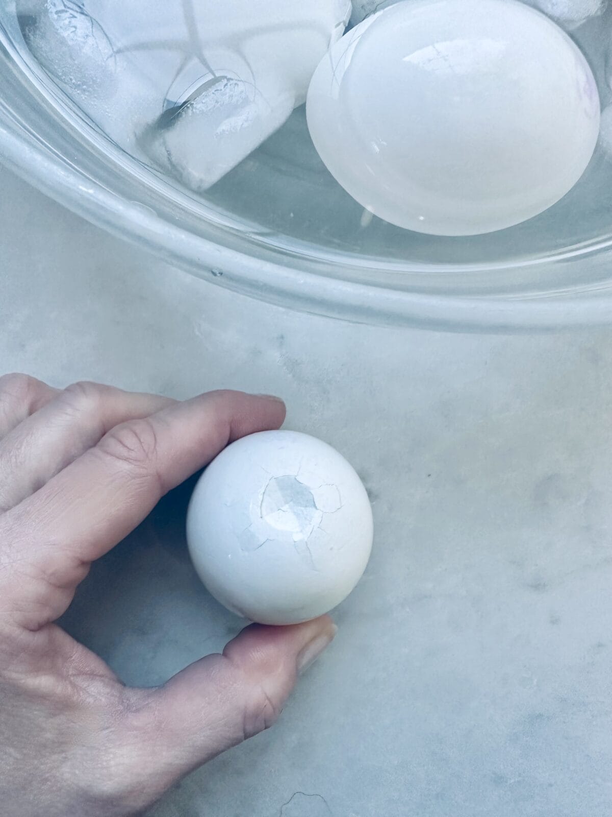 A hand holds a hard-boiled egg with small cracks in the shell, while another peeled egg and ice cubes are visible in a glass bowl on a light surface.